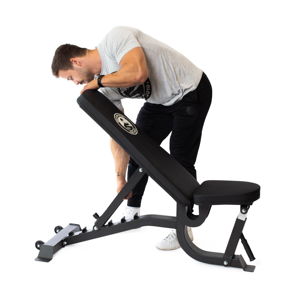 A man in a gray t-shirt and black pants adjusts a Bells of Steel Adjustable Utility Bench featuring a black padded seat and backrest. This bench, capable of multiple incline angles, is set against a white background.