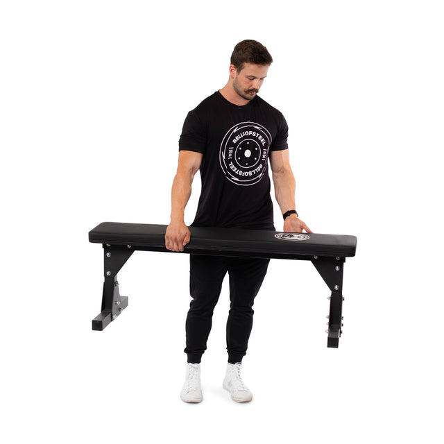 A man in black attire lifts one end of the Bells of Steel Flat Utility Bench, showcasing its 661lb max capacity. Set against a plain white background, this budget-friendly bench is an ideal gym addition.