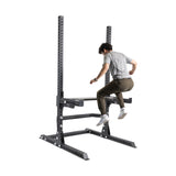 A person in a gray shirt and brown pants performs a box jump on the Bells of Steel Utility Seat for Hydra/Manticore. This robust exercise rack resembles a weight bench and features customizable holes, all set against a plain white background.