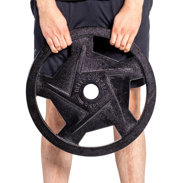 A person holds a Black Mighty Grip Olympic Weight Plate by Bells of Steel, featuring a star design and precision grip handles for ergonomics. They wear dark shorts against a plain white background.