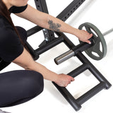 A person with a floral forearm tattoo loads a weight plate onto an Olympic bar secured to a squat rack, next to the Bells of Steel Viking Press Attachment on the gym floor.