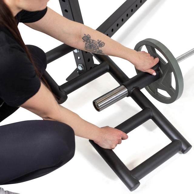 A person with a floral forearm tattoo loads a weight plate onto an Olympic bar secured to a squat rack, next to the Bells of Steel Viking Press Attachment on the gym floor.