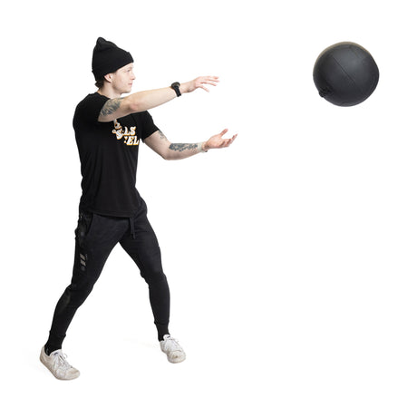 Against a white background, a person in black beanie, t-shirt, and pants reaches out with legs apart to catch a Bells of Steel Triple Stitched Medicine Ball.