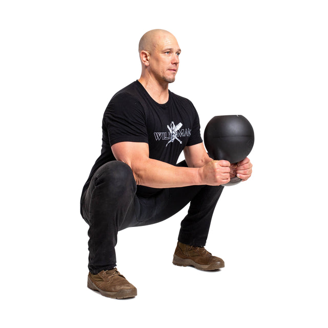 A person squats with the Mark Wildman Adjustable Kettlebell by Bells of Steel, wearing a black T-shirt, dark pants, and Wildman Athletica's brown shoes against a plain white backdrop.