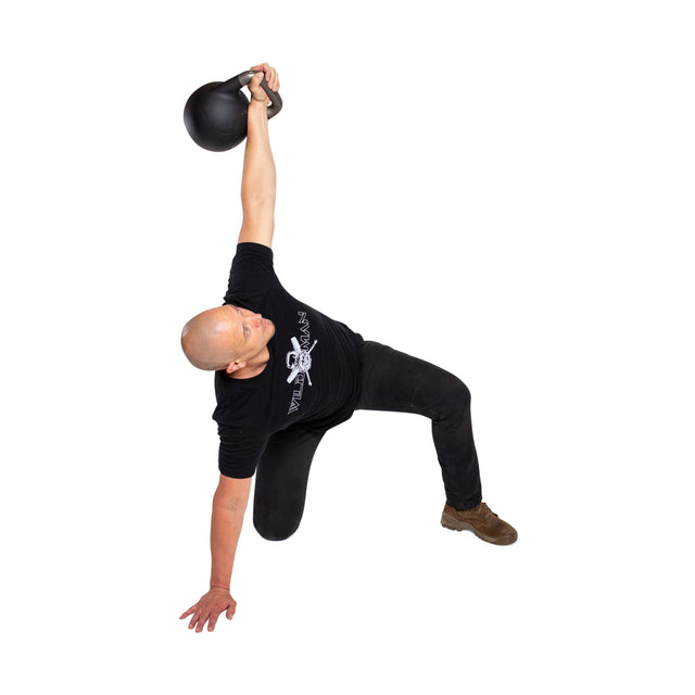 A person expertly performs a Turkish get-up using the Mark Wildman Adjustable Kettlebell by Bells of Steel. They balance on one knee with the opposite hand grounded, dressed in a sleek black shirt and pants against a stark white backdrop.