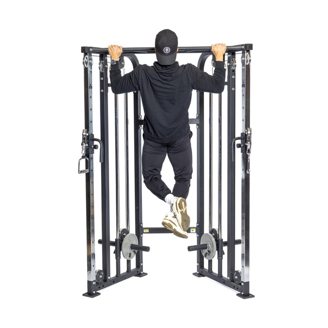 A person in black athletic clothing and a cap performs a pull-up on a Bells of Steel Functional Trainer, facing away from the camera. They use cable attachments with bent knees, and weights are visible on the machine.