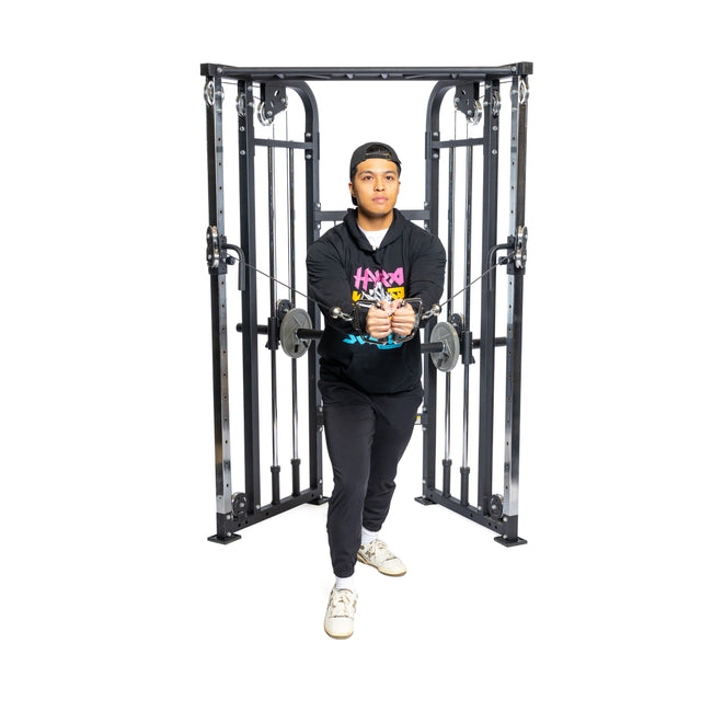 A person in a black hoodie and pants works out on a Bells of Steel Functional Trainer, skillfully using the cable attachments with both hands while standing with one foot slightly forward against a white background.