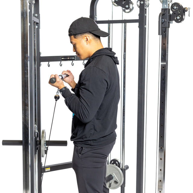 A person in a black hoodie and cap uses a Bells of Steel Functional Trainer for exercise, performing a bicep curl with the cable attachments in a gym filled with various equipment.