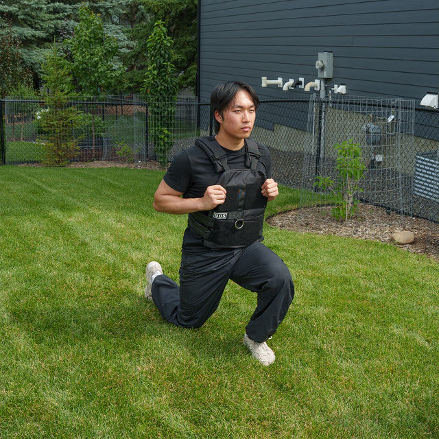 A person wearing the Bells of Steel USA Weighted Vest and Plates over black workout clothes performs a lunge on green grass in a fenced backyard beside a modern dark house.
