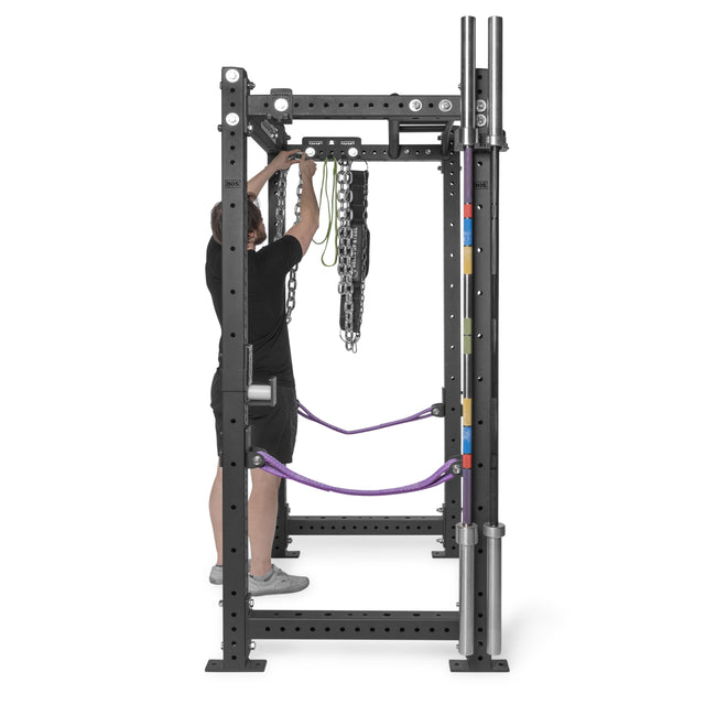 A person in athletic wear adjusts lifting chains on a black metal rack equipped with the Bells of Steel Accessory Holder (Wall or Rack Mounted), along with purple resistance bands and color-coded barbell collars, viewed from the front.