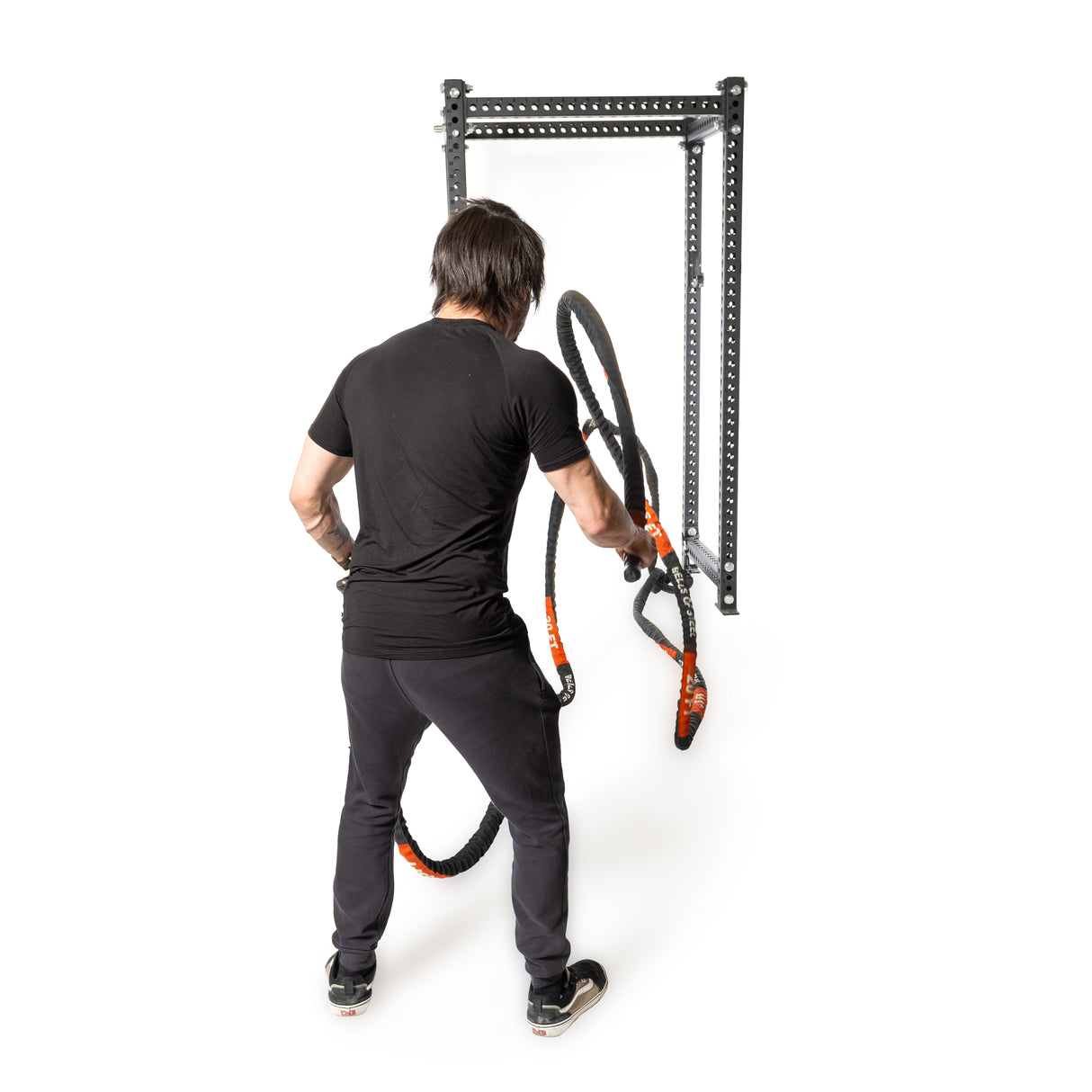 A person in black workout attire vigorously uses the Bells of Steel Battle Rope, featuring a durable sleeve, attached to a metal wall bracket. Facing away from the camera, they are focused on a conditioning workout against a plain white background.