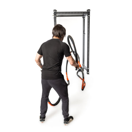 A person in black workout attire vigorously uses the Bells of Steel Battle Rope, featuring a durable sleeve, attached to a metal wall bracket. Facing away from the camera, they are focused on a conditioning workout against a plain white background.