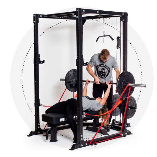 Using the Home Gym Builder, a woman bench presses with resistance bands in a power rack, aided by a man. Both wear gray shirts, using a bench and extra weights on the barbell.