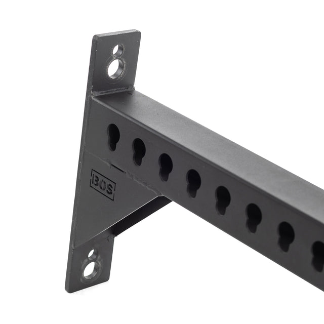 Close-up of the Bells of Steel Utility Arm, a sturdy black metal bracket perfect for wall studs or power racks. It includes multiple keyhole slots, two round mounting holes on one end, and a rectangular cutout displaying "BOS.