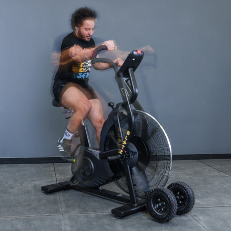 A man in black workout gear cycles vigorously on the Bells of Steel Blitz Max Air Bike, his motion blurred as he demonstrates the intensity and performance of this elite commercial fitness equipment.