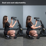 A woman demonstrates dual-axis seat adjustability on the Bells of Steel Blitz Max Air Bike by changing both seat height and horizontal position. Text at the top reads, "Dual-axis seat adjustability," against a plain gray wall background.
