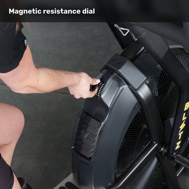 A person adjusts the resistance dial on a Bells of Steel Blitz Max Air Bike, commercial fitness equipment with a large fan and sturdy frame.