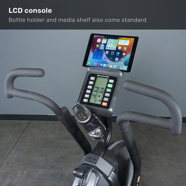 The Bells of Steel Blitz Max Air Bike offers a display console, media shelf for an iPad, bottle holder, and dual handlebars—ideal for commercial gyms—all showcased against a gray floor and wall backdrop.
