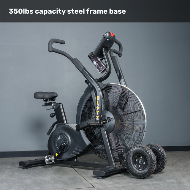 The Bells of Steel Blitz Max Air Bike is a black commercial fitness bike with a large fan wheel, steel frame (350lbs capacity), and digital display. Two spare spoked wheels are beside it.