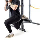 Wearing checkered slip-ons and white socks, a person performs a Bulgarian split squat using a dumbbell with the Split Squat Leg Roller Rack Attachment from Bells of Steel. The padded support adds comfort, while orange bands and black clothing complete the setup next to a power rack attachment.