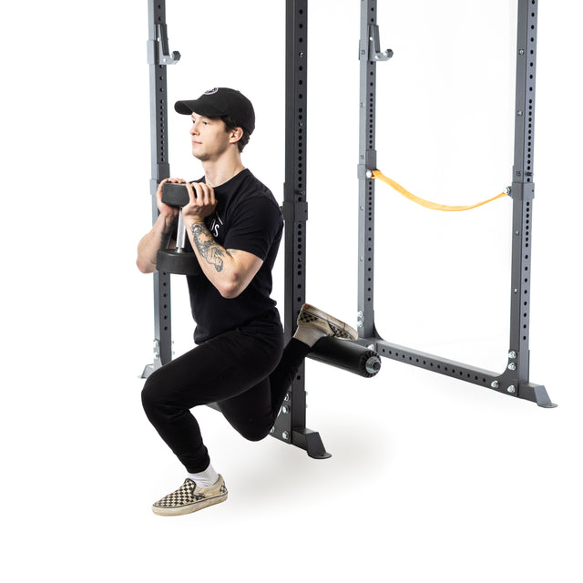 Dressed in black workout clothes and a cap, a person performs a Bulgarian split squat with a dumbbell using the Bells of Steel Split Squat Leg Roller Rack Attachment in front of a power rack, set against a white gym background.