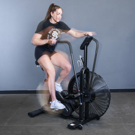 A woman in athletic wear rides the Bells of Steel Blitz Air Bike indoors, her arms and legs blurred in motion against a gray wall and tiled floor.