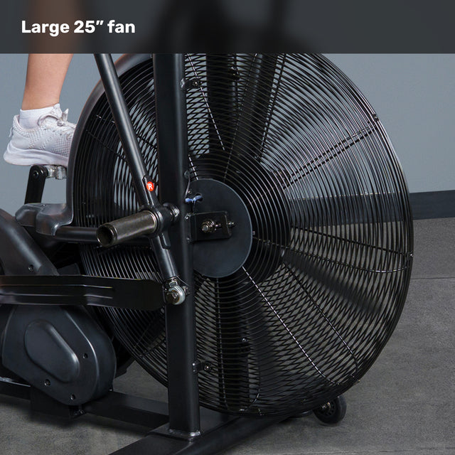 Close-up of the 25-inch fan on the Blitz Air Bike by Bells of Steel, showing a user's lower leg and white sneaker on the pedal. A gray wall and floor in the background emphasize its sturdy commercial-grade design.