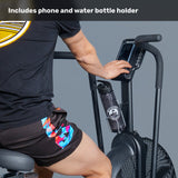 A person sits on the Bells of Steel Blitz Air Bike, pointing at its display. A water bottle is in the built-in holder. The image text says, "Includes phone and water bottle holder.