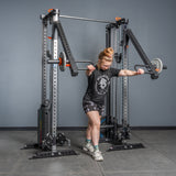 Wearing athletic gear, a person uses the Bells of Steel Lever Arms Rack Attachment—loaded with Olympic plates—on a squat rack. The gym has gray walls/flooring, an adjustable pull-up bar, and other fitness equipment in view.
