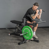In a gym, a man wearing athletic gear does preacher curls on a machine with green weight plates. Nearby is the Bells of Steel USA Legacy Leg Extension / Hamstring Curl Machine - Plate Loaded, while he focuses on training his arms.