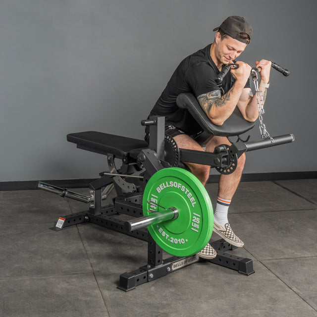 In a gym, a man wearing athletic gear does preacher curls on a machine with green weight plates. Nearby is the Bells of Steel USA Legacy Leg Extension / Hamstring Curl Machine - Plate Loaded, while he focuses on training his arms.