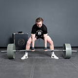 A man in a black shirt and shorts prepares to lift the Bells of Steel USA Deadlift Bar in a gym, standing on a gray floor with various equipment and a black box behind him.
