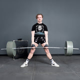 Wearing a black t-shirt, shorts, and white trainers, a person performs a sumo deadlift with the Bells of Steel USA Deadlift Bar in a gym with gray walls and flooring, demonstrating impressive powerlifting strength.