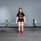 A woman in glasses, a black tee, patterned shorts, and red socks powerlifts with the Bells of Steel USA Deadlift Bar in a gym featuring gray walls and flooring.