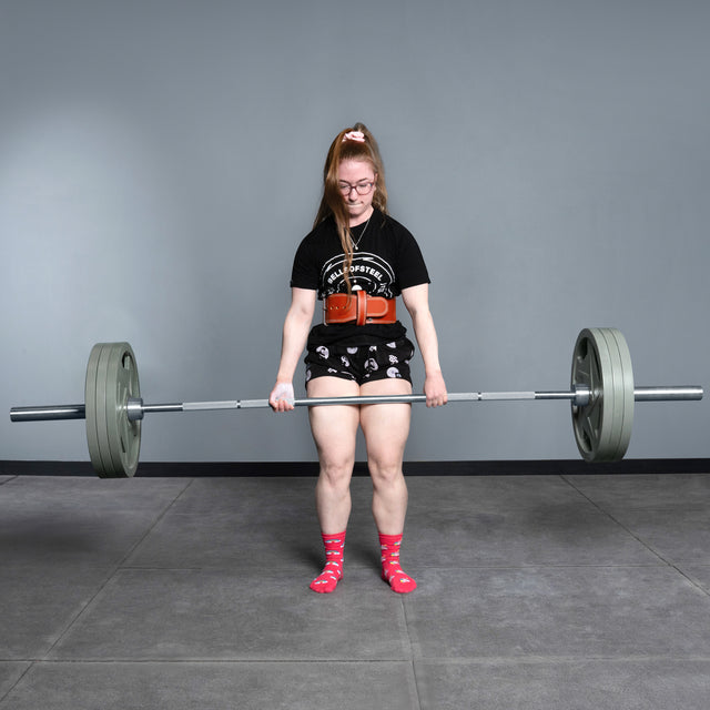A woman in glasses, a black tee, patterned shorts, and red socks powerlifts with the Bells of Steel USA Deadlift Bar in a gym featuring gray walls and flooring.