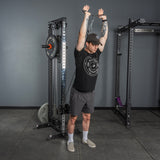 Wearing a black t-shirt, shorts, and cap, a person stands in the gym using the Bells of Steel USA Flat-Iron Tricep Press Cable Attachment for overhead triceps extensions, arms extended to maximize tricep activation.