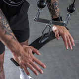 A person with tattooed arms grips the Bells of Steel USA Flat-Iron Tricep Press Cable Attachment in a gym, ready to work out. They're dressed in black shorts, white socks, and athletic shoes.