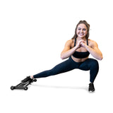A woman in athletic wear demonstrates a hamstring exercise with a smile, performing a side lunge using the Bells of Steel Glute Ham Slider against a white background, hands clasped at her chest.