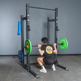 A person in a black t-shirt and shorts performs a barbell squat with green and blue plates on a Bells of Steel Hydra Half Rack Builder (3" x 3", ⅝" holes), with a blue towel hanging on the rack in a home gym.