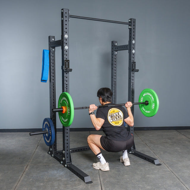 A person in a black t-shirt and shorts performs a barbell squat with green and blue plates on a Bells of Steel Hydra Half Rack Builder (3" x 3", ⅝" holes), with a blue towel hanging on the rack in a home gym.