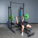 A person in black is bench pressing a barbell with green and blue plates in a Bells of Steel Hydra Half Rack Builder (3" x 3", ⅝" Holes) inside a gym.