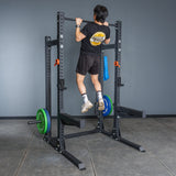 A person in a black t-shirt and shorts performs a pull-up on the Bells of Steel Hydra Half Rack Builder (3" x 3", ⅝" holes) in a home gym with gray walls and flooring, weight plates loaded for an ideal workout setup.