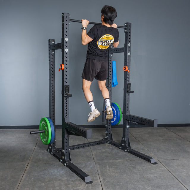 A person in a black t-shirt and shorts performs a pull-up on the Bells of Steel Hydra Half Rack Builder (3" x 3", ⅝" holes) in a home gym with gray walls and flooring, weight plates loaded for an ideal workout setup.