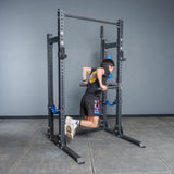 A person in athletic gear does a dip on parallel bars attached to the Bells of Steel Hydra Half Rack Builder (3" x 3", ⅝" Holes) in a home gym with a gray wall and tiled floor.