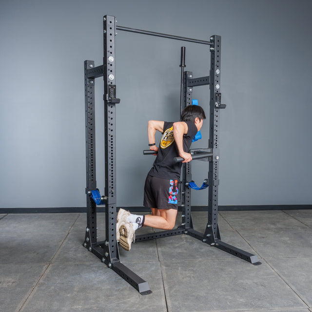 A person in athletic gear does a dip on parallel bars attached to the Bells of Steel Hydra Half Rack Builder (3" x 3", ⅝" Holes) in a home gym with a gray wall and tiled floor.