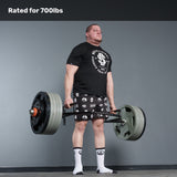 A man in a black shirt and patterned shorts deadlifts a heavy barbell with the Bells of Steel Open Trap Bar / Hex Bar, rated for 700 lbs, in a gym. He wears white socks, standing on a gray floor against a gray wall, highlighting intense strength training.