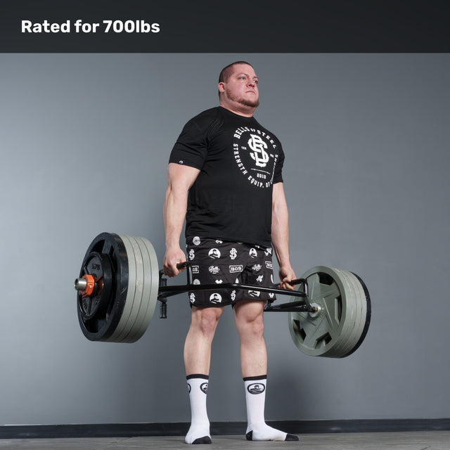 A man in a black shirt and patterned shorts deadlifts a heavy barbell with the Bells of Steel Open Trap Bar / Hex Bar, rated for 700 lbs, in a gym. He wears white socks, standing on a gray floor against a gray wall, highlighting intense strength training.