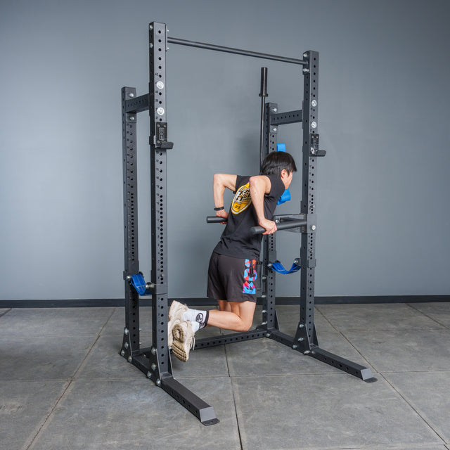 A person in a black shirt and shorts does dip exercises on parallel bars attached to the Bells of Steel Hydra Half Rack - Prebuilt (3" x 3", ⅝" Holes) in a home gym with gray walls and a tiled floor.