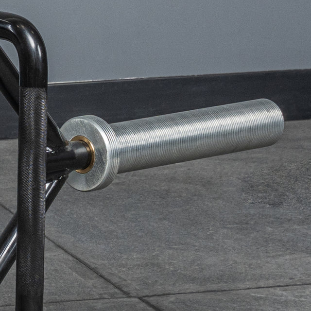 A close-up of the sleeve and handle of the Bells of Steel Open Trap Bar / Hex Bar resting on a gray gym floor, with a gray wall and black baseboard in the background.