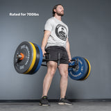 A man lifts the Bells of Steel Open Trap Bar / Hex Bar with large weight plates in a gym. He wears a grey t-shirt, black shorts, and black sneakers. Text on image: "Rated for 700lbs.
